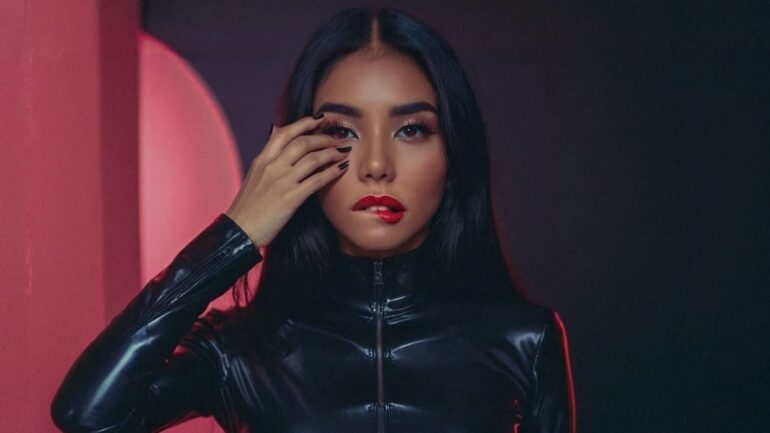 Beautiful woman with long black hair and red lipstick, wearing a black plastic-like outfit, standing in front of a pink backdrop.