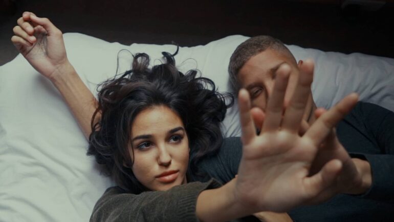 A close-up of a young woman lying on a bed, reaching out her hand, with a man lying beside her, both engaged in an intimate moment.