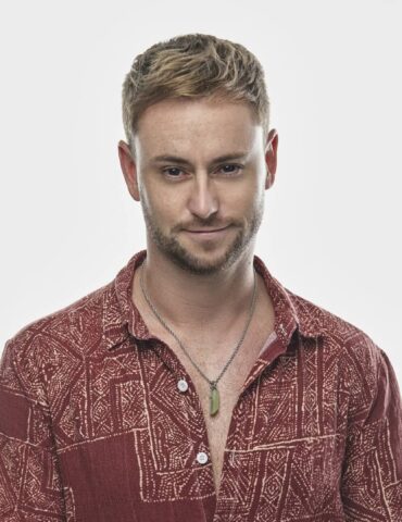 Kenn Colt in a red patterned shirt, hands folded, with a reflective smile, expressing the emotion behind his music.